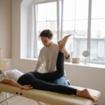Massage therapist provides leg therapy to client on a table in a sunlit room.